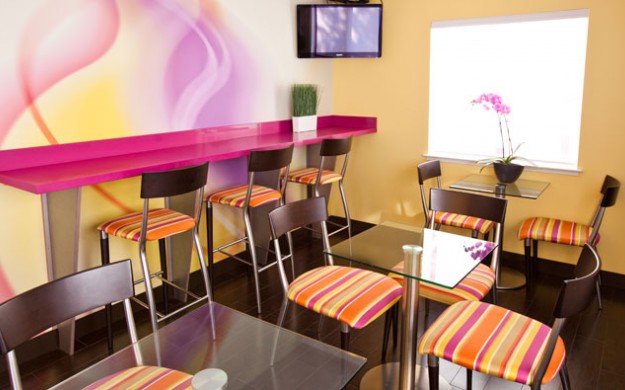 dining area contains small glass tables with 2 chairs and a wall of cafe bar seating. The room is decorated with bright wall paper, chairs and accents.