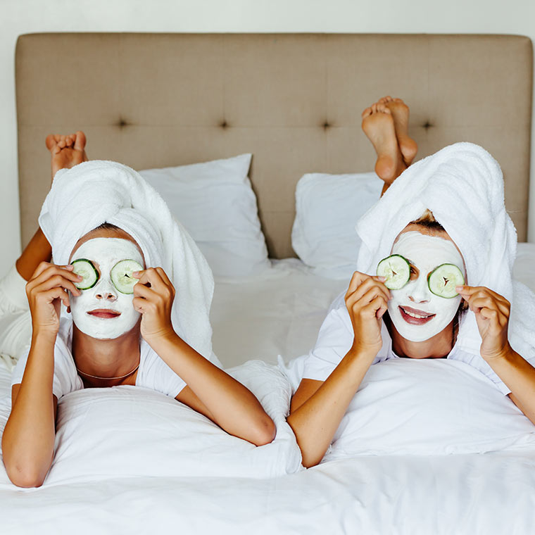 two women laying in bed wearing face masks and holding cucumber slices up to their eyes
