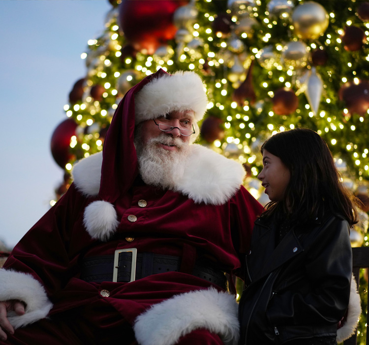 little girl standing next to santa claus with a giant light up and decorated christmas tree