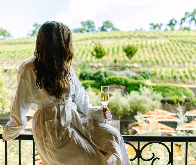 Woman wearing a robe drinking champagne on a balcony overlooking a vineyard