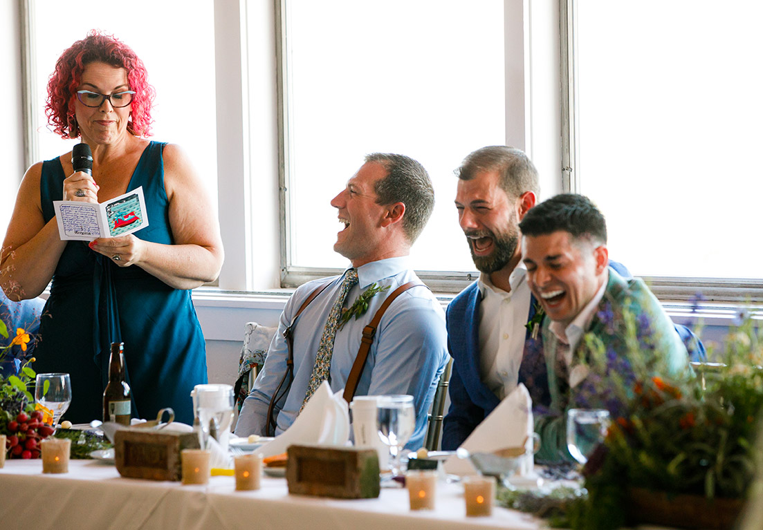 woman with pink hair standing up with a microphone next to three men who are sitting and laughing