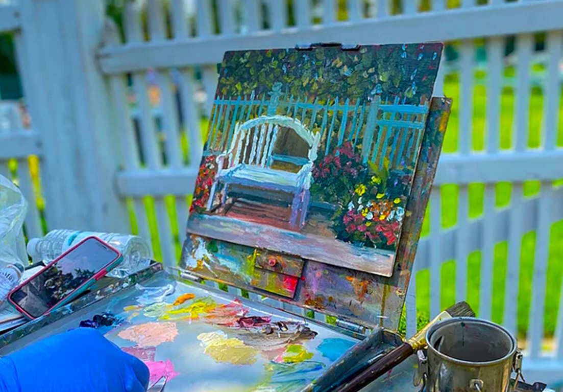 a hand painting a picture outside in the grass area facing a white fence