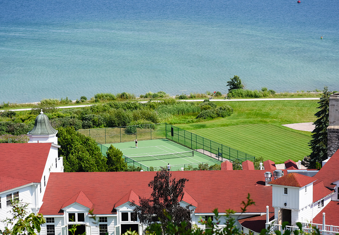 over view of the tennis court near the beach surrounded by green grass and trees all around with a walk way between the green grass and beach