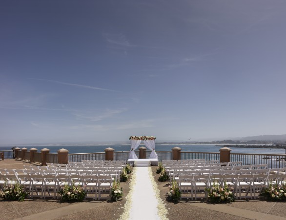 wedding ceremony overlooking the monterey bay with two sections of white chairs separated by a path of white rose pedals leading to a white curtain altar