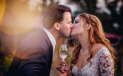 a bride and groom kissing while holding glasses of rose as the sun is setting