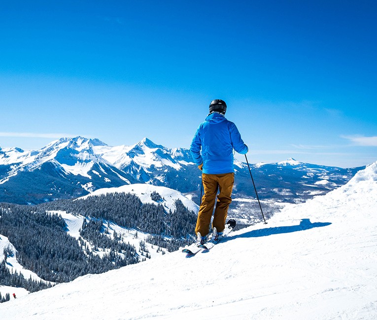 a skier wearing a blue jacket and looking at the snow covered mountains and clear blue skies