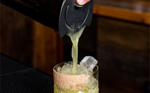a bartender pouring a light green beverage into a rocks glass 
