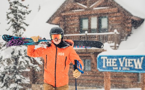a skier wearing a bright orange jacket and holding his gear over his shoulder while in front of the view bar and grill