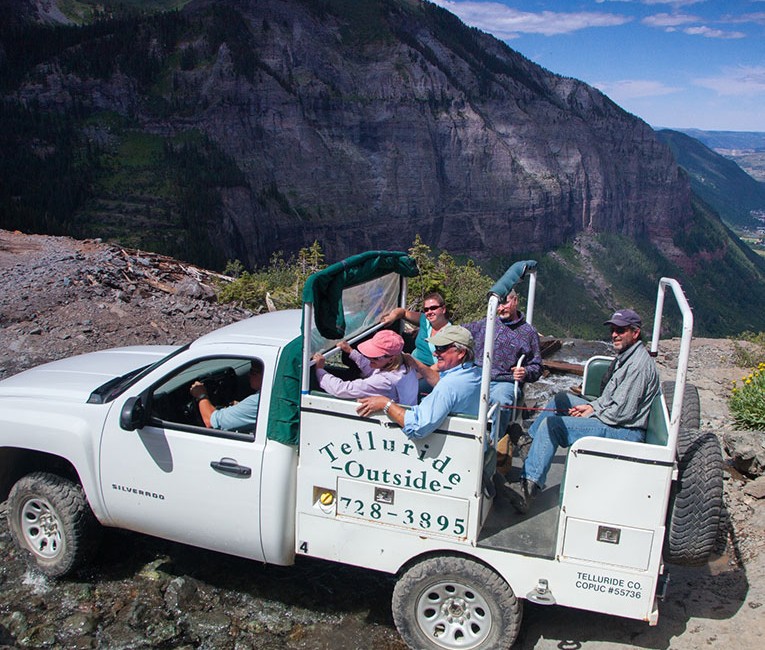 a white truck driving off road with people sitting in the back and mountains in the distance