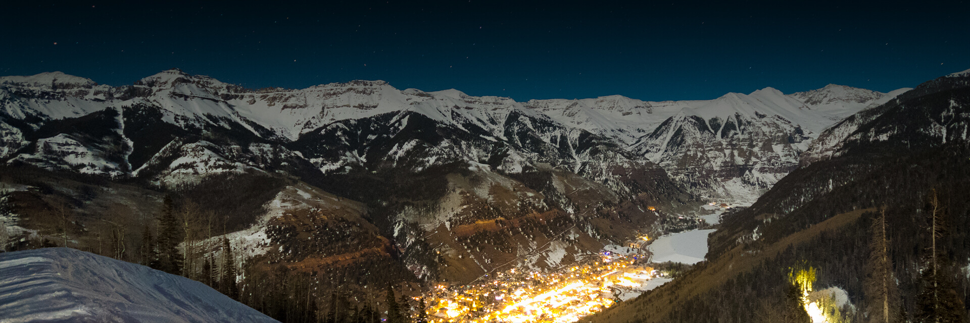the town with lots of lights at night with snow covered mountains in the distance