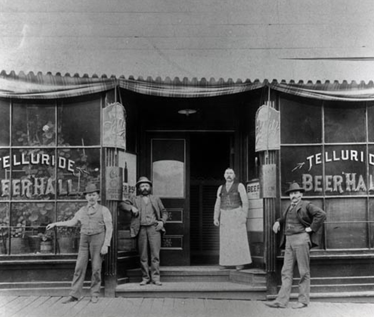 a black and white photo of men standing in front of an old fashioned store