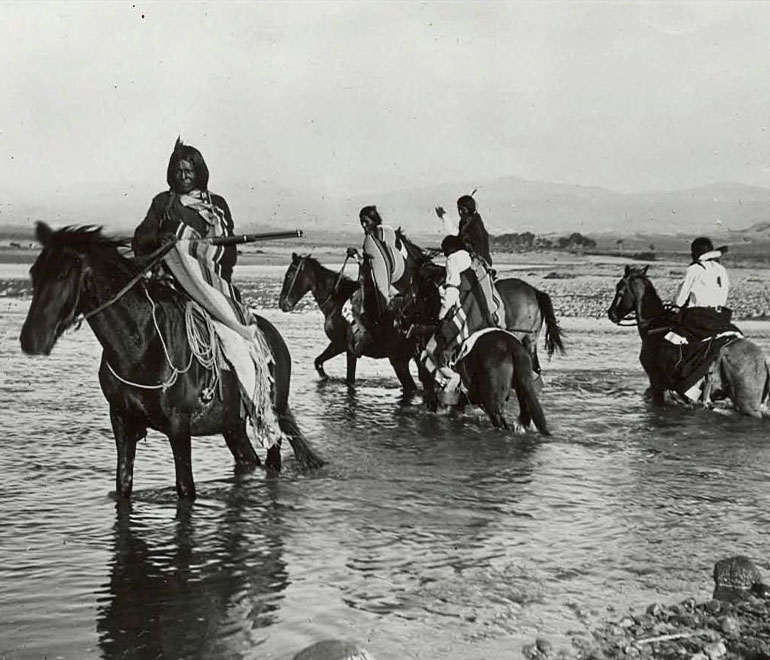 a black and white image of native americans riding horses through the shallow water