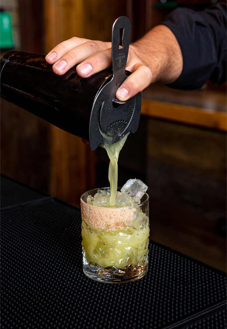 a bartender pouring a light green beverage into a rocks glass 