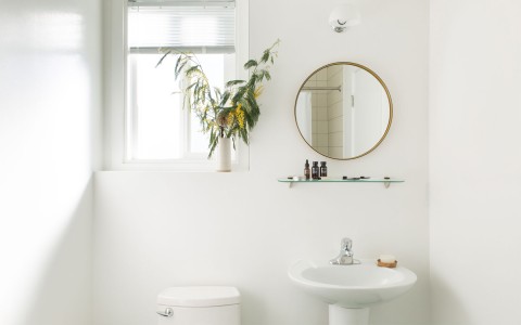 all white bathroom with pedistal sink, mirror and glass shelf. Next to toilet and small window above
