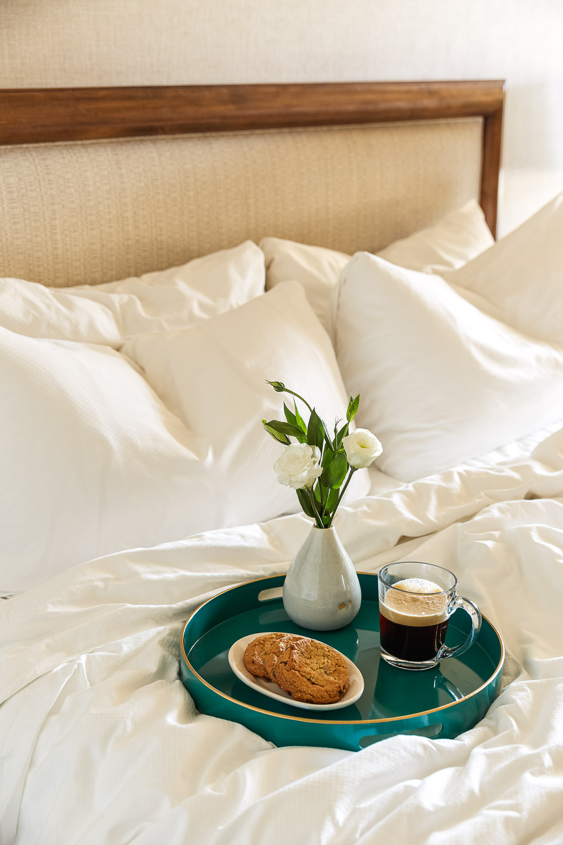 Front view of a white bed, with a tray on top, a coffee cup a biscuit on a plate and a flower vase 