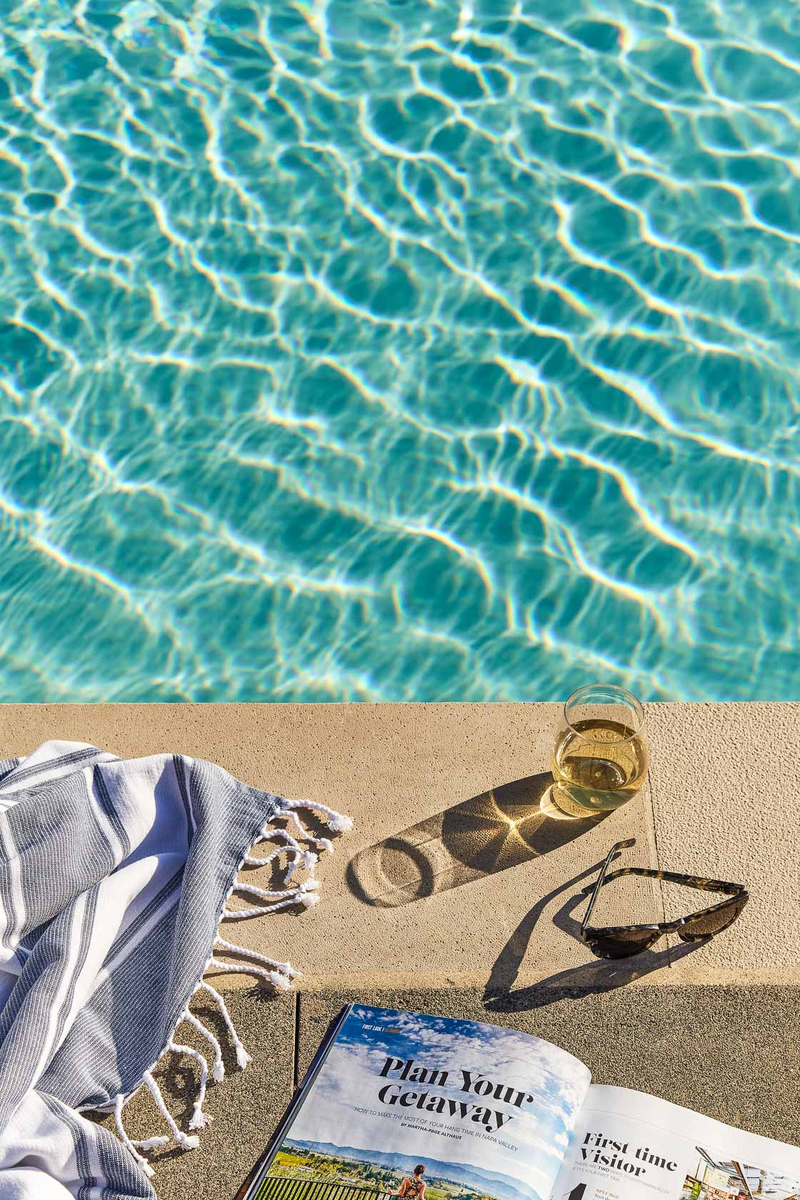 View of a part of a pool with a beverage, a pair of sunglasses, a magazine and a beach sarong