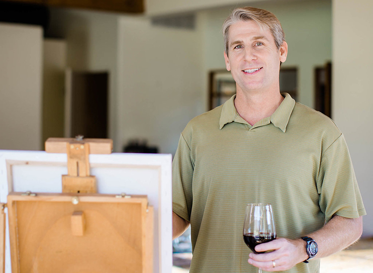 man holding glass of wine next to easel