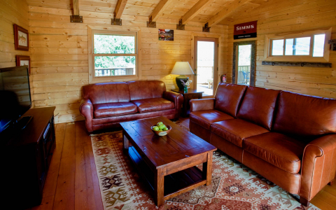 Log cabin interior with a leather couch and love seat, wooden table and entertainment center with a flatscreen TV