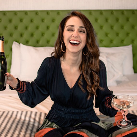 woman sitting on her hotel bed with champagne and laughing