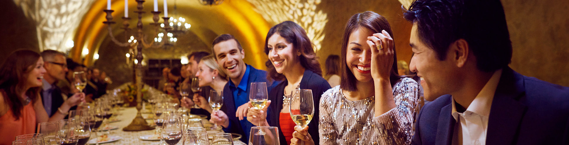 People gathered sitting and smiling  in a fancy large restaurant table having a glass of white wine