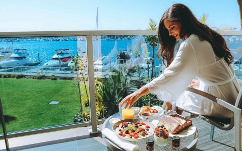 Woman sitting on her balcony on a white robe with her breakfast served on the table next to her and the marina in the back on a sunny day