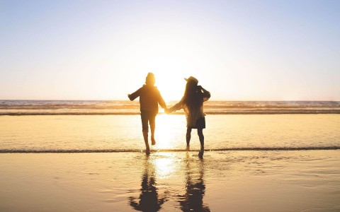 Couple holding hands with their feet on the ocean walking towards the sunset 