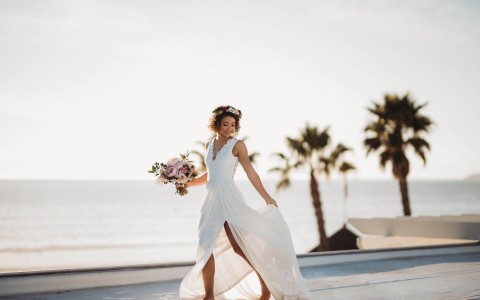 Bride walking near the beach holding her dress with the left hand and a floral arrangement on the right hand on a beautiful sunset