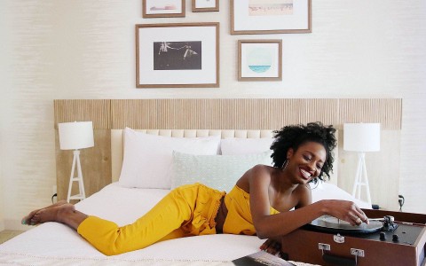 Black woman on a yellow suit smiling while lying down the bed playing a song on a turntable