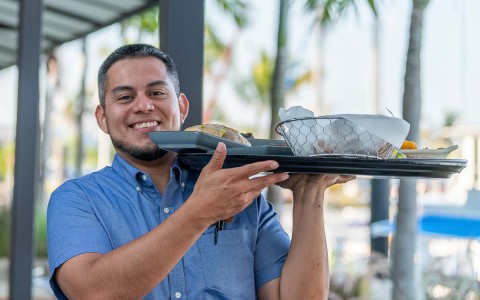 waiter guy smiling looking straight to the camera while he is carrying a big tray with food 