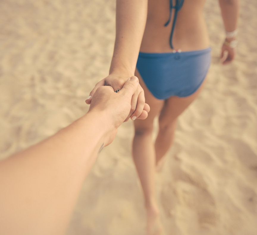 woman in bikini leading male hand down the beach