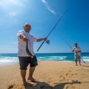 two men with fishing poles on the beach and one guy holding a fish