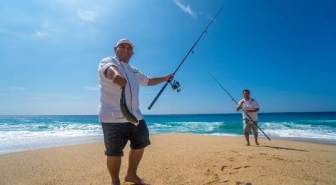 two men with fishing poles on the beach and one guy holding a fish 