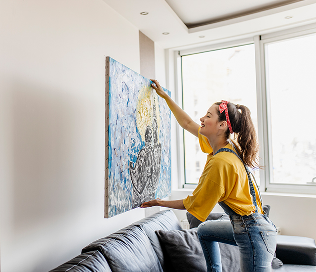woman with yellow shirt and jean overall hanging a painting in the wall 