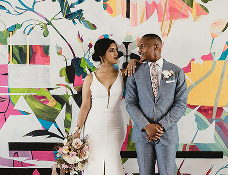 A married couple looking at each other and standing on a colorful wall