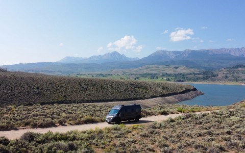 a distance shot of the van driving down a pebbled path, on a clear sunny day, surrounded by hills and a bright blue lake