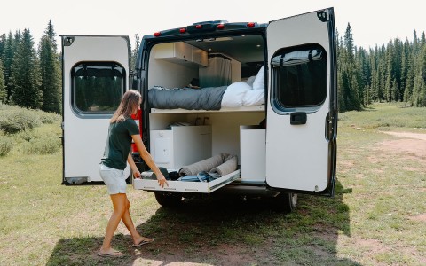 Back view of a Roameo Van, the back doors are open and there is a clear view of the bed on the top half and a lady pulling out a drawer from the bottom filled with belongings and amenities. 