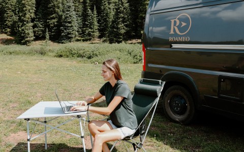 A Roameo customer sitting outside on a sunny day, next to the Roameo van. On a pull out chair & desk working on their computer.