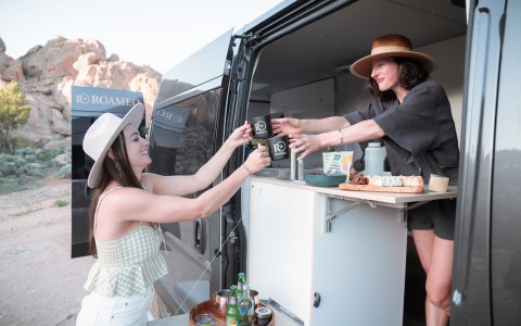 A woman in handing another woman out of the van a cup of coffee