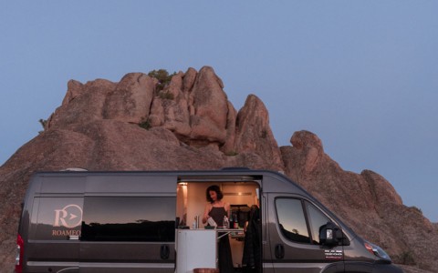 A van parked infront of a large rock and a woman cooking in the van