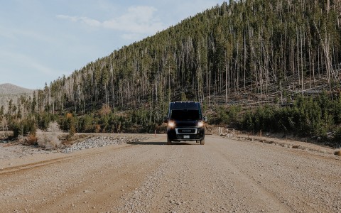 roameo van from the front, on a dirt and rocky road with tall thing trees in the back on a curve from a mountain hill