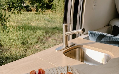 a set up of crackers and fruit on the little table connecting to the sink with the green trees in the background with the van door open