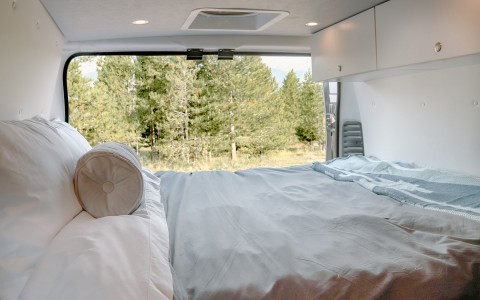 view of the outdoor green trees from the bed, the back doors of the van are open. white sheets, two pillows and a gray comforter 