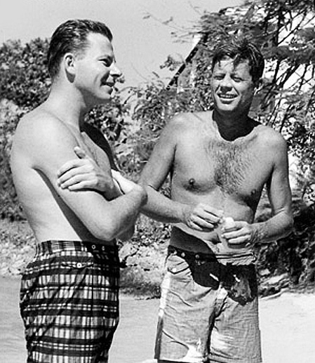 JFK standing on a beach with swim trunks chatting with a man