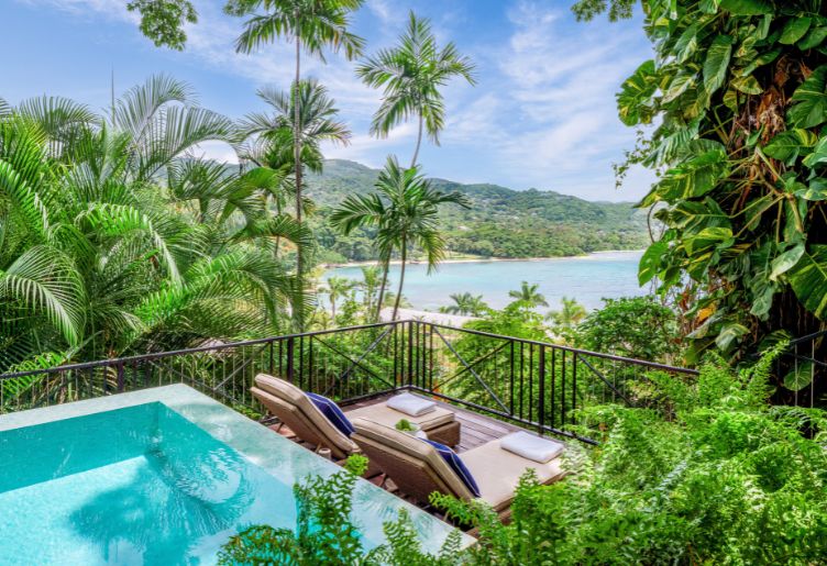 view of a private pool and seating from a villa overlooking a beach and green foliage