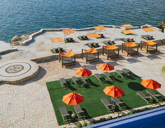 view of reclined patio loungers and cabanas with orange umbrellas between a pool and ocean