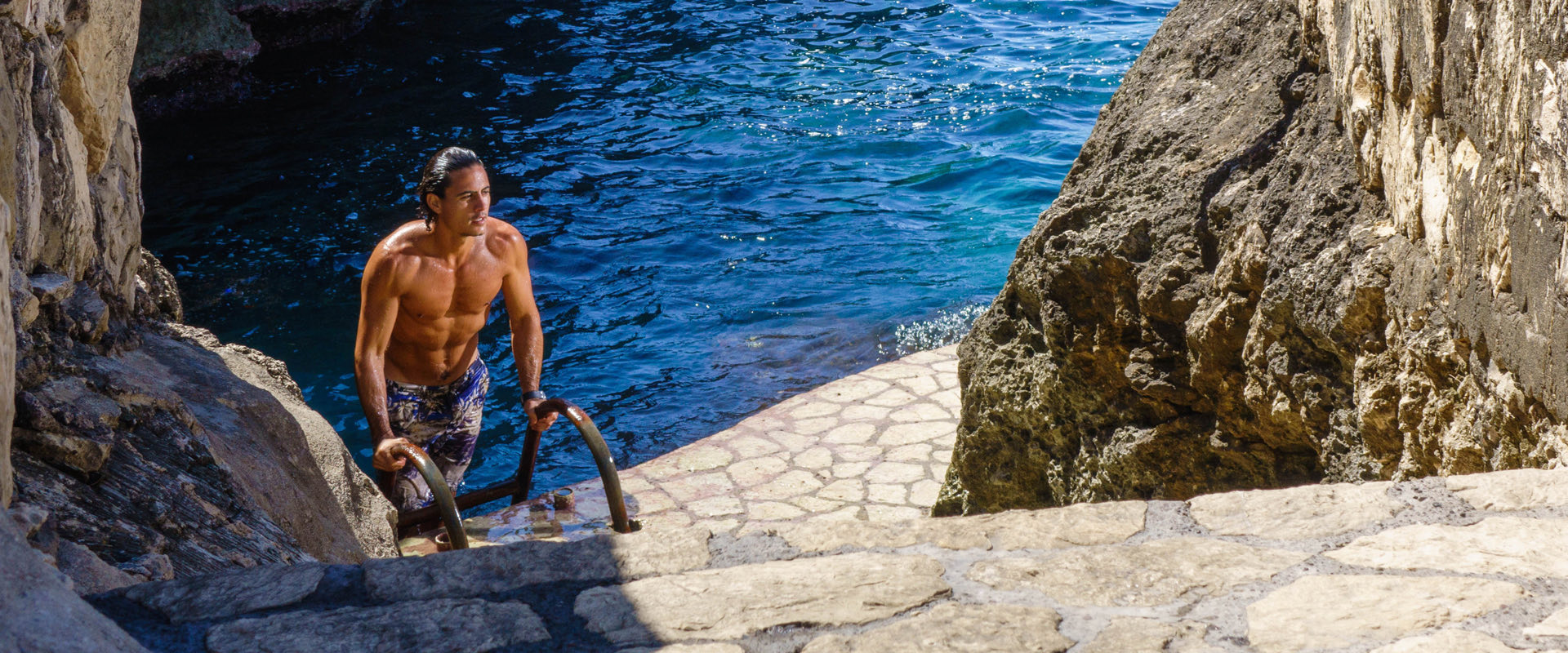 athletic man in swim trunks climbing up ladder up cliff from ocean