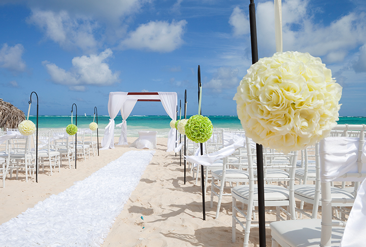 wedding aisle runner on the sand lined with chairs leading to a simple wooden arch with draped with white fabric, ocean view in background