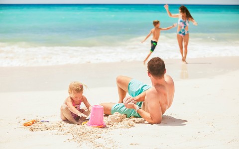 man building sandcastle with daughter and watching mom and son play near ocean