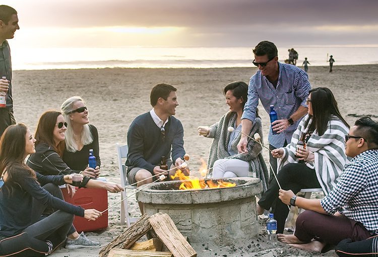 Group of friends sitting around fire pit with ocean in the back