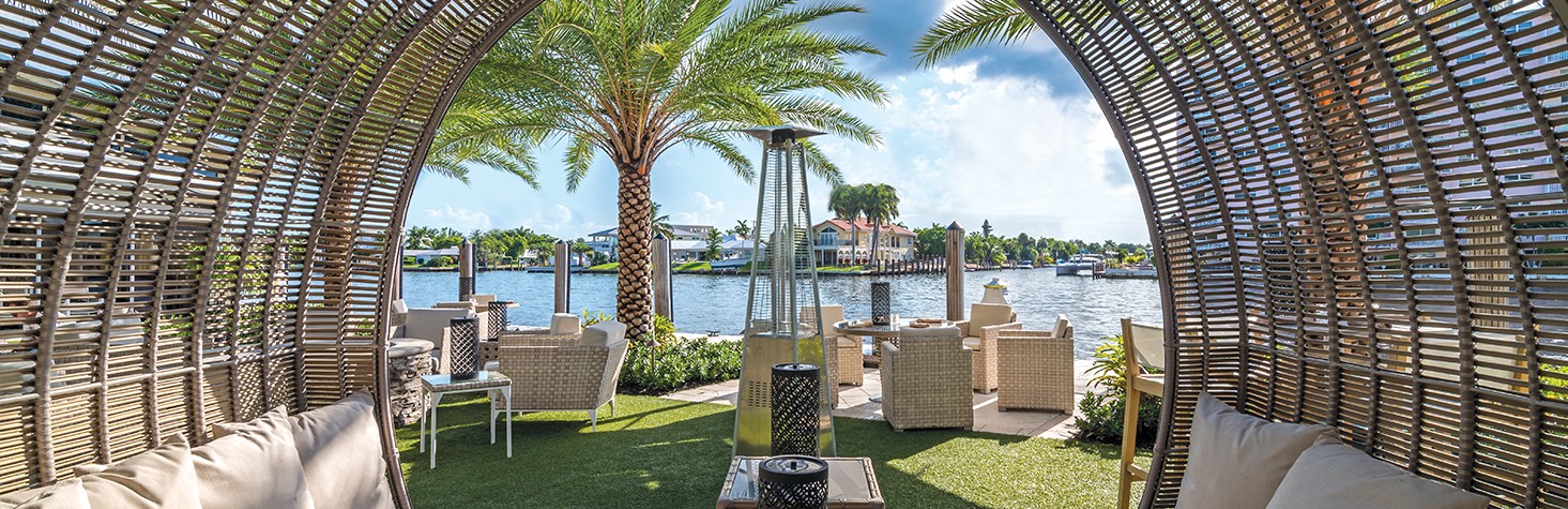 A circular wicker seating area at the outdoor grass patio next to the water with additional chairs and tables 